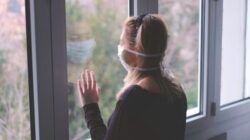 admin-ajax (4) Woman with mask staring out window dealing with isolation during covid pandemic