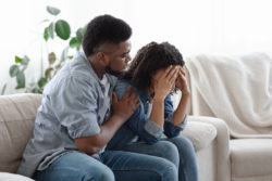 Dependent Relationship Concept. Young Black Guy Soothing His Depressed Girlfriend At Home, Embracing Her On Couch In Living Room