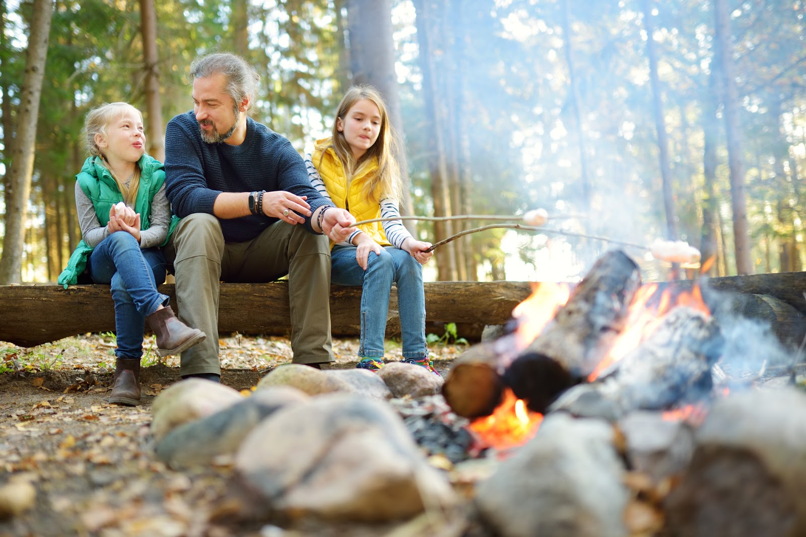 A father with his two daughters sitting by a campfire in the woods roasting marshmallows