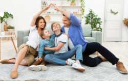 Housing for family. Young parents joining hands making symbolic house roof sitting with children on floor in new apartment.