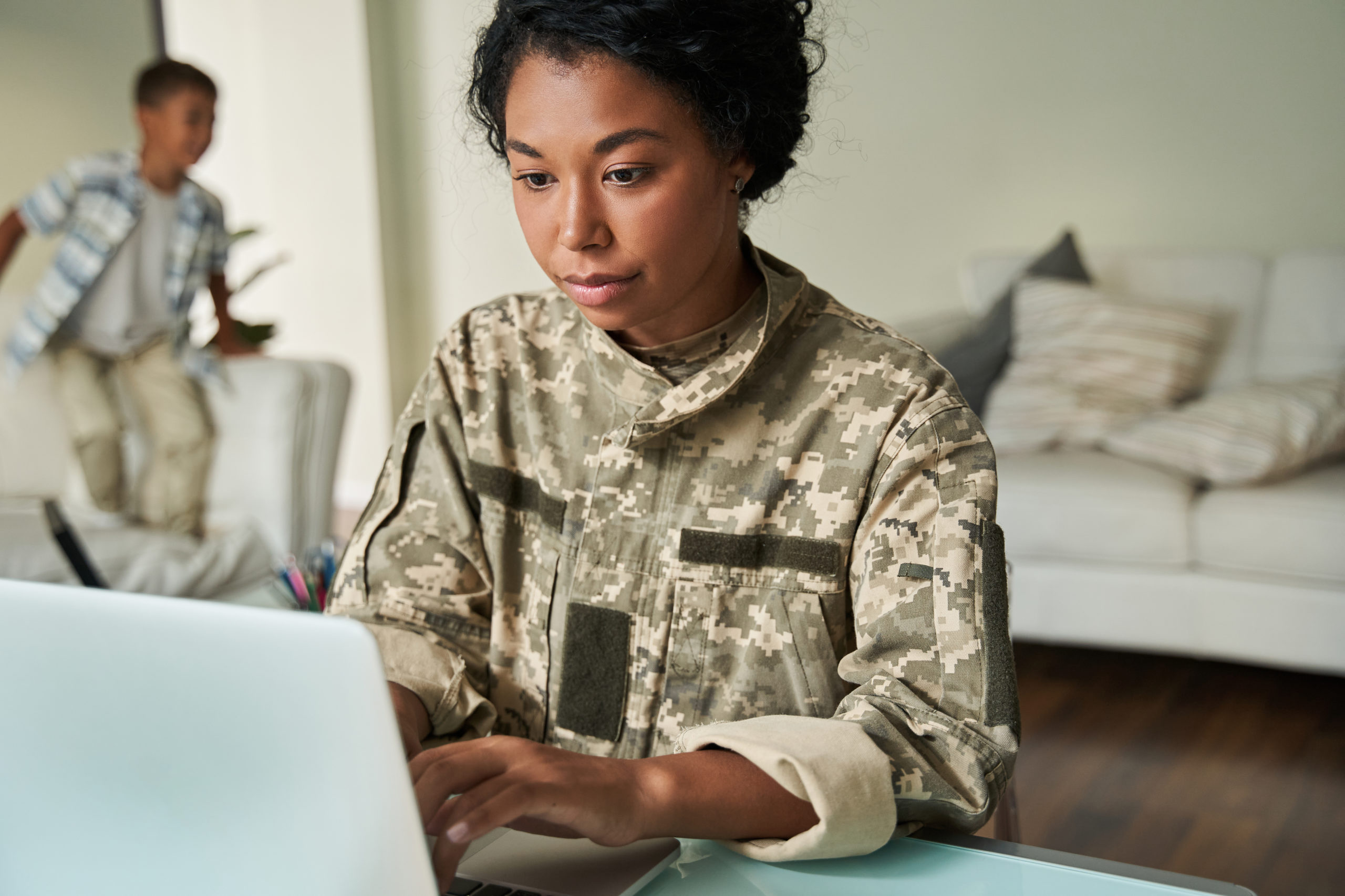 Focused black woman using laptop at table. Concept of modern woman. Female soldier wear camouflage uniform. After war rehabilitation. Blurred background of little boy. Interior of modern apartment