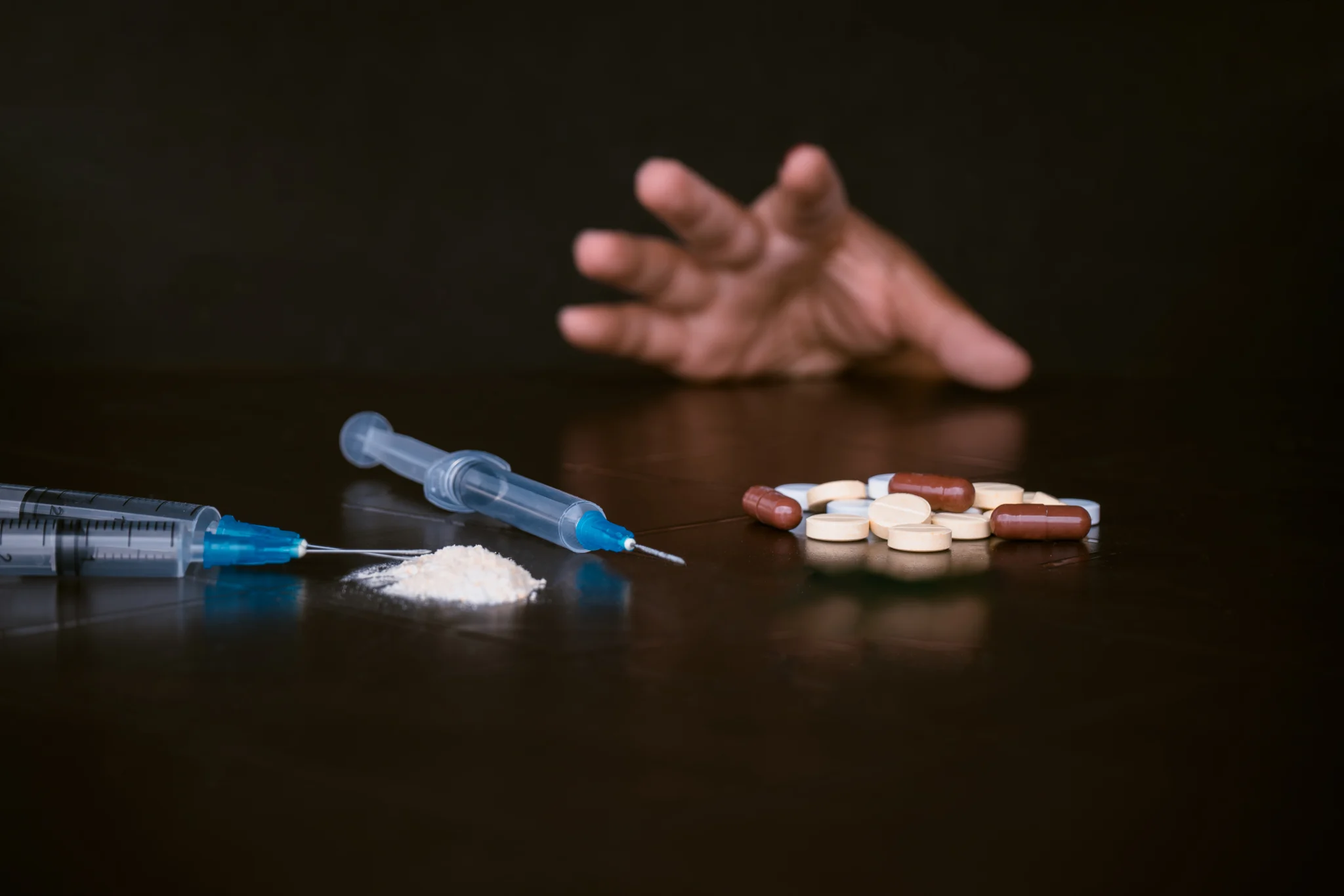 Needles laying on table next to pile of white powder and a pile of tablets next to them with a hand in the background reaching towards the drugs