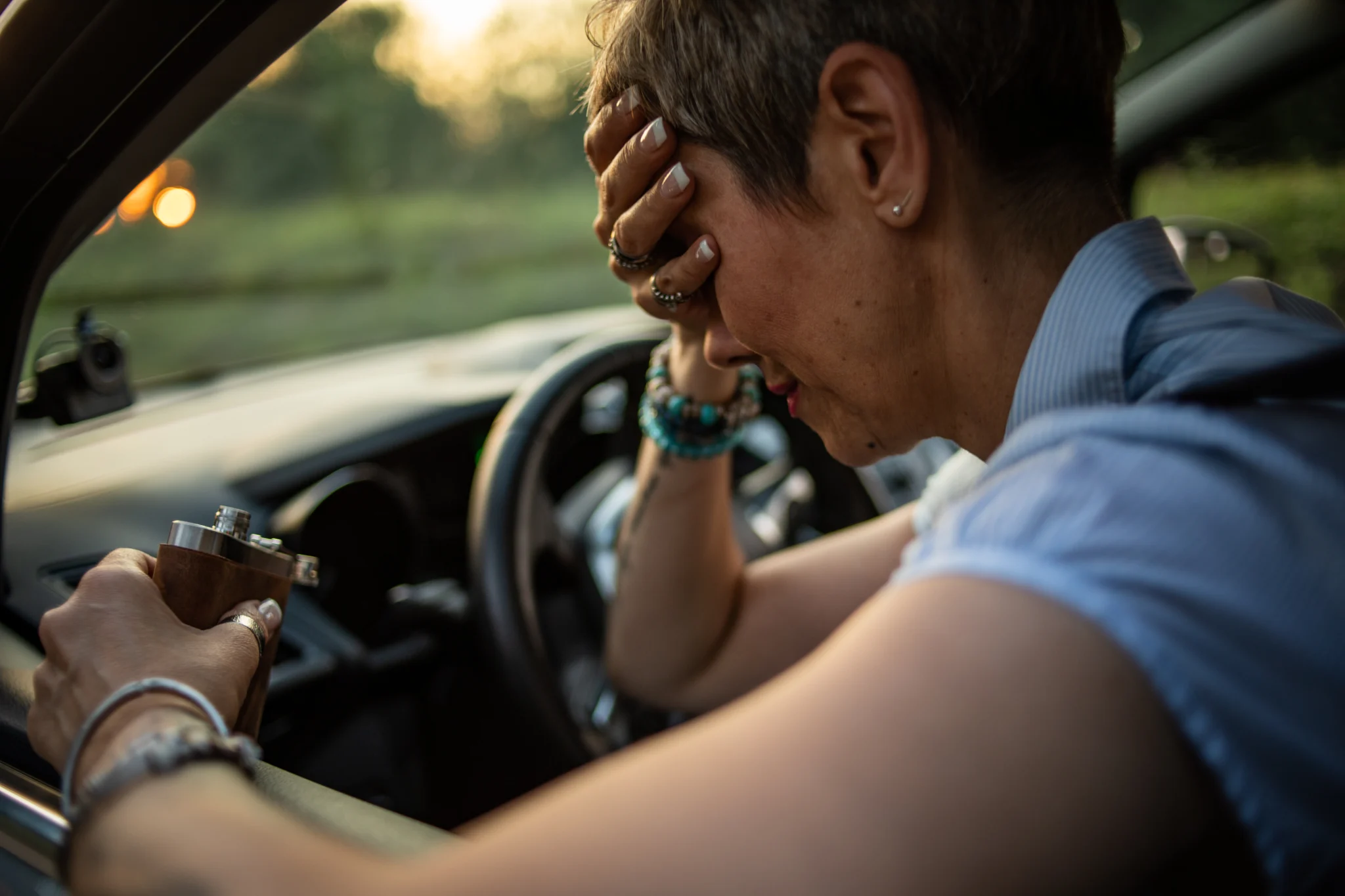 A mature woman sits behind the wheel and drinks brandy struggling to find alcohol addiction treatment