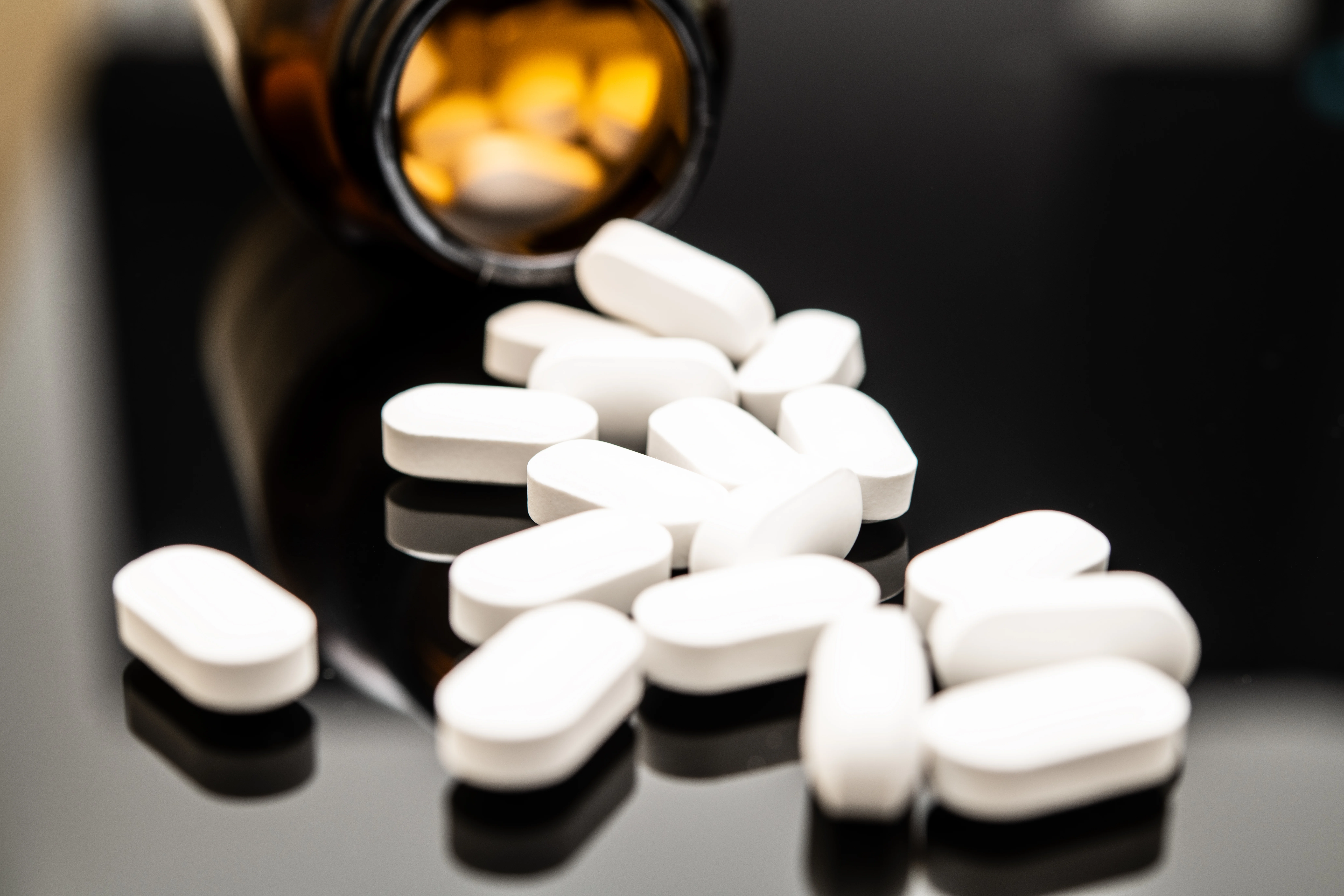 Close-up image of white opioid pills spilled from a dark amber prescription bottle onto a reflective surface, highlighting the dangers of opioid overdose and misuse.