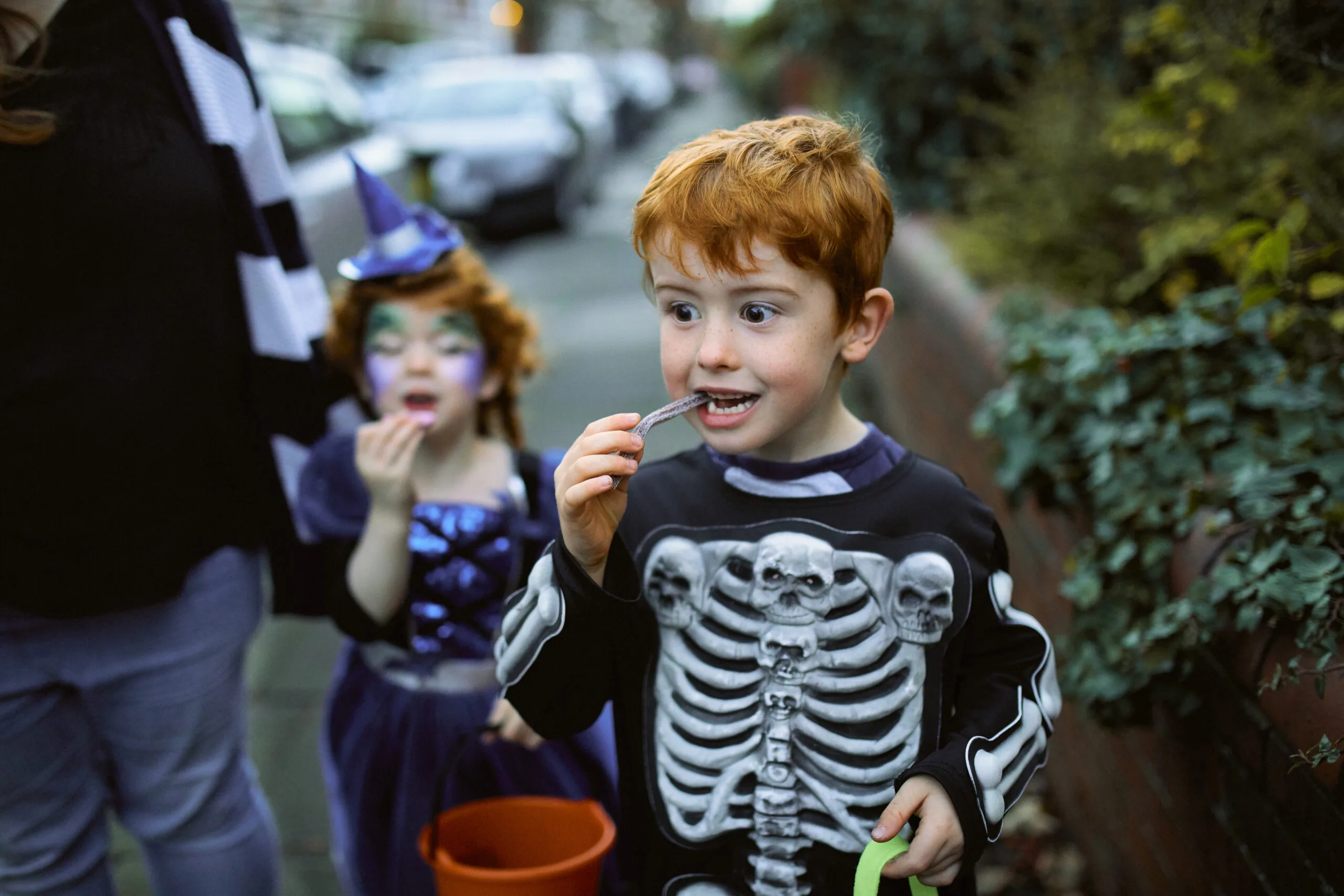 Little boy eating halloween candy while trick or treating in suburban streets with his mother and sister.