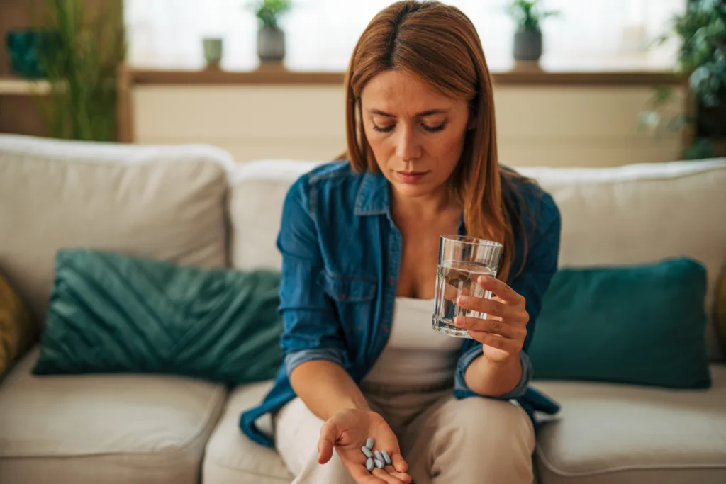 Mid adult woman sitting on a sofa at home, taking pills with water, feeling unwell and grappling with the challenges of illness and discomfort in her daily life