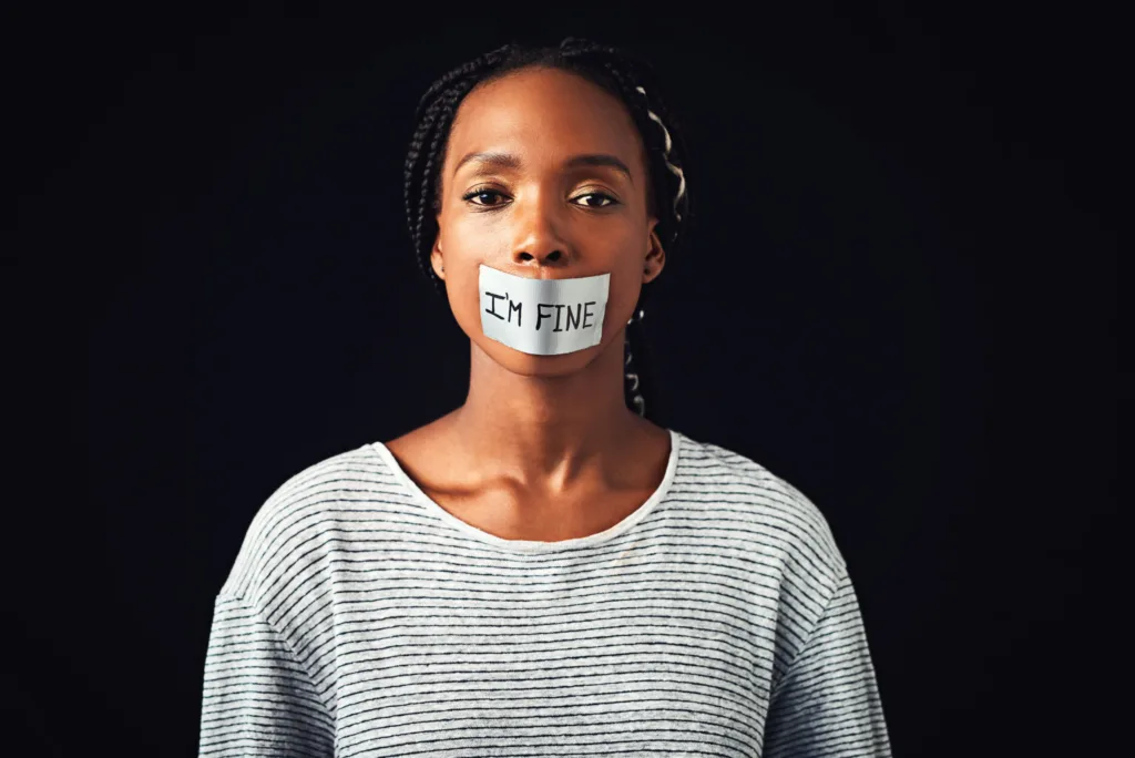 Studio shot of a young woman with a label saying “I’m fine” covering her mouth against a black background