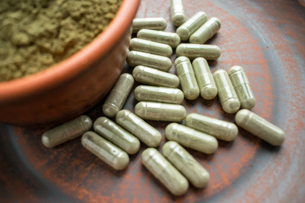 Supplement kratom green capsules and powder on brown plate. Herbal product alt-medicine kratom is  opioid. Home alternative pain remedy, opioid addiction, dangerous painkiller, overdose. Close up. Selective focus