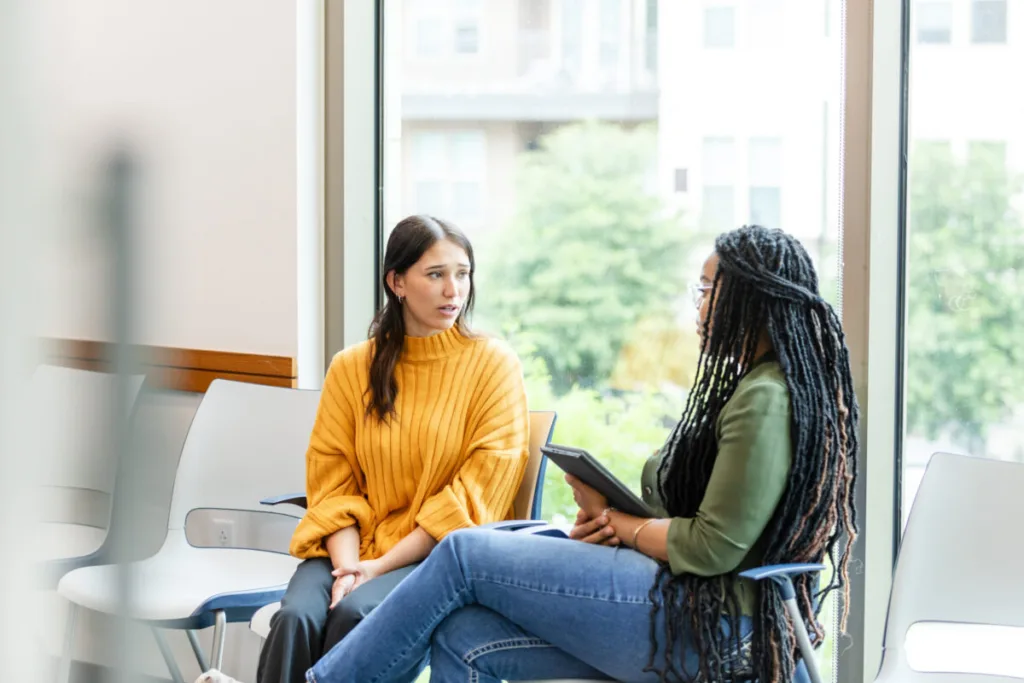 The young adult woman looks worried as she listens to the unrecognizable young adult female therapist.